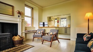 A living room with a fireplace, two armchairs, a sofa, and a kitchen area visible through an open door. - Peaceful Farmhouse, Kinclune Estate, Angus Glens (Kirkton of Kingoldrum)