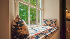 Un siège de fenêtre agrémenté d’un coussin à motifs et d’un oreiller, une vue sur la verdure à l’extérieur et une lampe sur la droite.