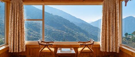A room with wooden walls and floor, a large window with curtains, two wooden chairs, and a table with a book.