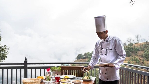 Un chef sirve comida en un balcón con vistas a un paisaje montañoso.