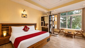 Una habitación de hotel con una cama grande, mesitas de noche, un escritorio con sillas, una estantería y una ventana con vistas a árboles.