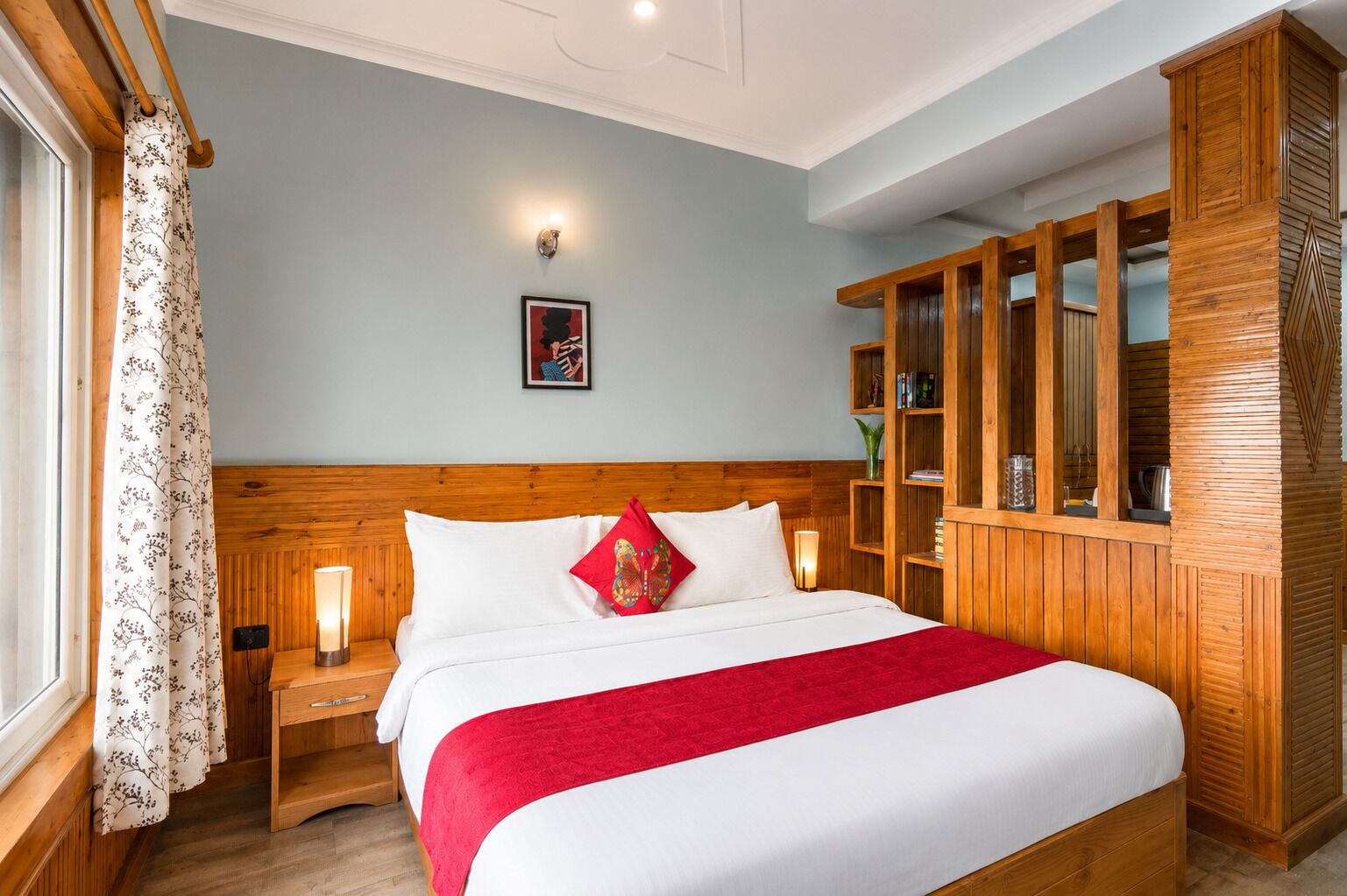 A hotel room with a wooden headboard, a large bed with white bedding and a red throw, a nightstand with a lamp, a wooden wardrobe, and a framed picture on the wall.