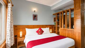 A hotel room with a wooden headboard, a large bed with white bedding and a red throw, a nightstand with a lamp, a wooden wardrobe, and a framed picture on the wall.