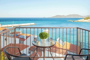 A balcony with a table, chairs, and a potted plant overlooking a coastal view with buildings and a pier. - Studio Tepes - Niki Samos Sea View Balcony Suite (Kokkari)