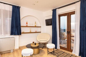 A modern living room with a round table, two white stools, a television, and a balcony with a view. - Studio Tepes - Natassa Samos SeaView Balcony Suite (Kokkari)