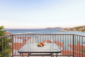 Ein Balkon mit Tisch für zwei Personen, der Blick auf die Küste mit Gebäuden und Booten bietet.