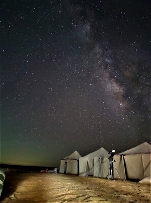 A night sky with visible stars and the Milky Way. There are tents and a tripod with a camera on the ground.