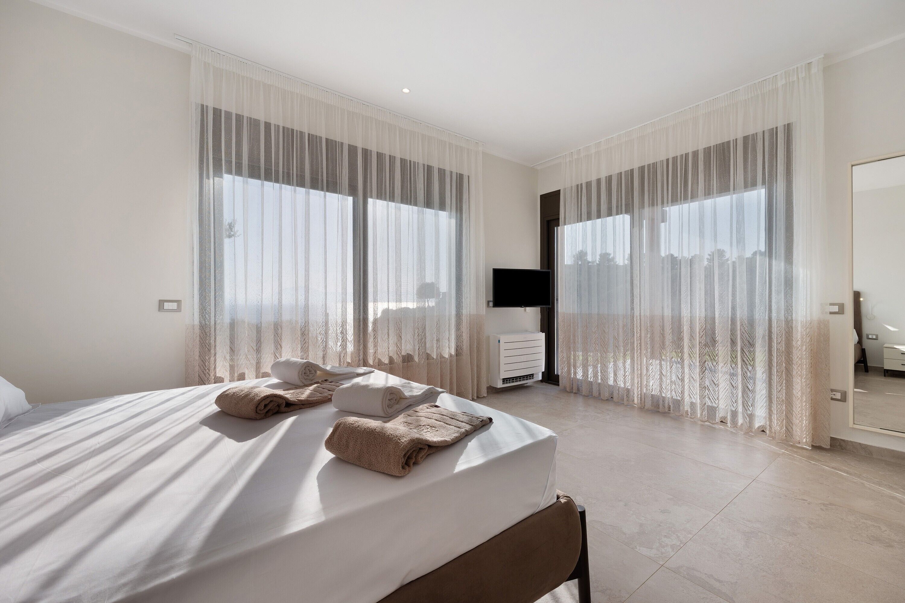 A modern hotel room with a large bed, a flat-screen TV, and a view of the city through sheer curtains.