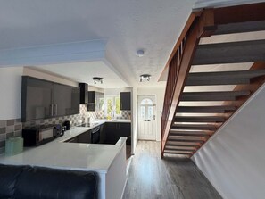 A modern kitchen with a central island, dark cabinetry, and a wooden staircase leading to another level. - 2BDR House with Private Garden and Parking (Greater London)