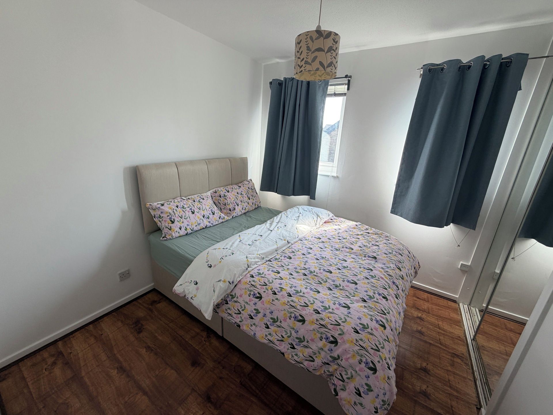 Un dormitorio con cama, ventana con cortinas, lámpara colgante y suelo de madera.