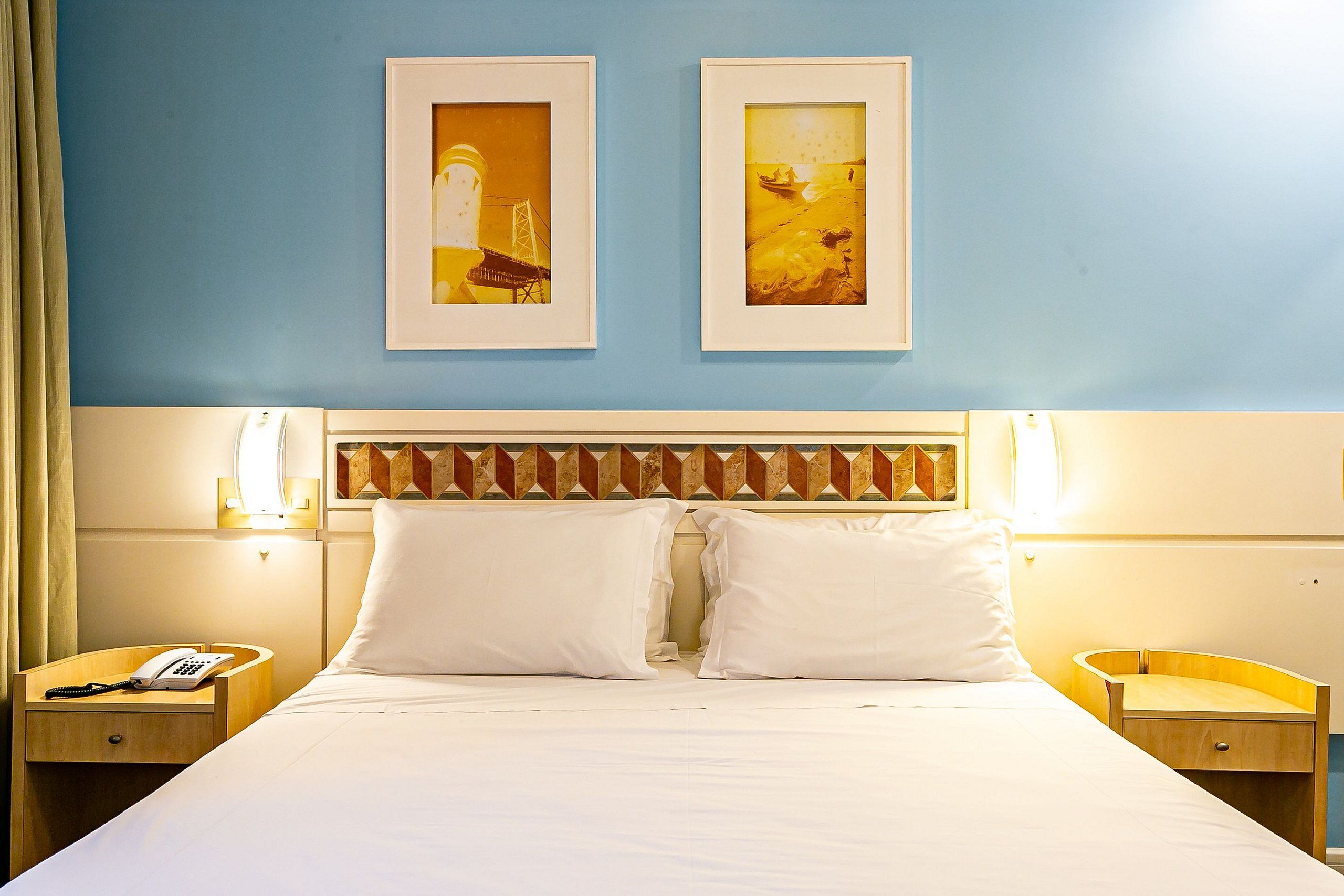 A hotel room with a large bed, two framed pictures on the wall, bedside lamps, a phone on the nightstand, and a small wooden side table.