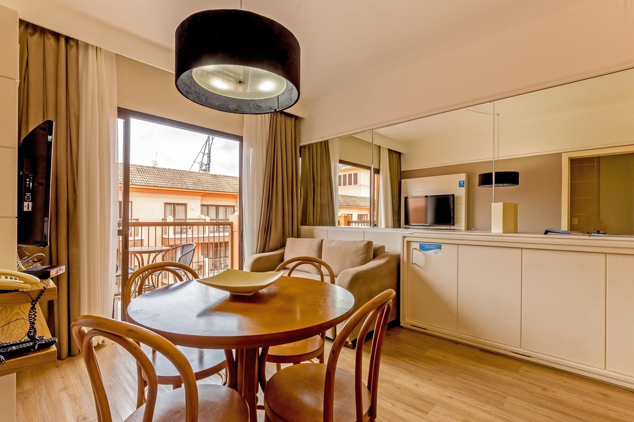 A modern hotel room with a dining area, a sofa, a TV, and a kitchenette.