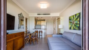A modern living room with a sofa, a dining area with chairs, a TV, a kitchen in the background, and a tropical artwork on the wall. - Oceanfront Suite in a Beachfront Resort Jbv122 (Florianópolis)