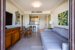 Ein modernes Wohnzimmer mit einer Couch, einem Essbereich mit Stühlen, einem Fernseher, einer Küche im Hintergrund und einem tropischen Kunstwerk an der Wand.