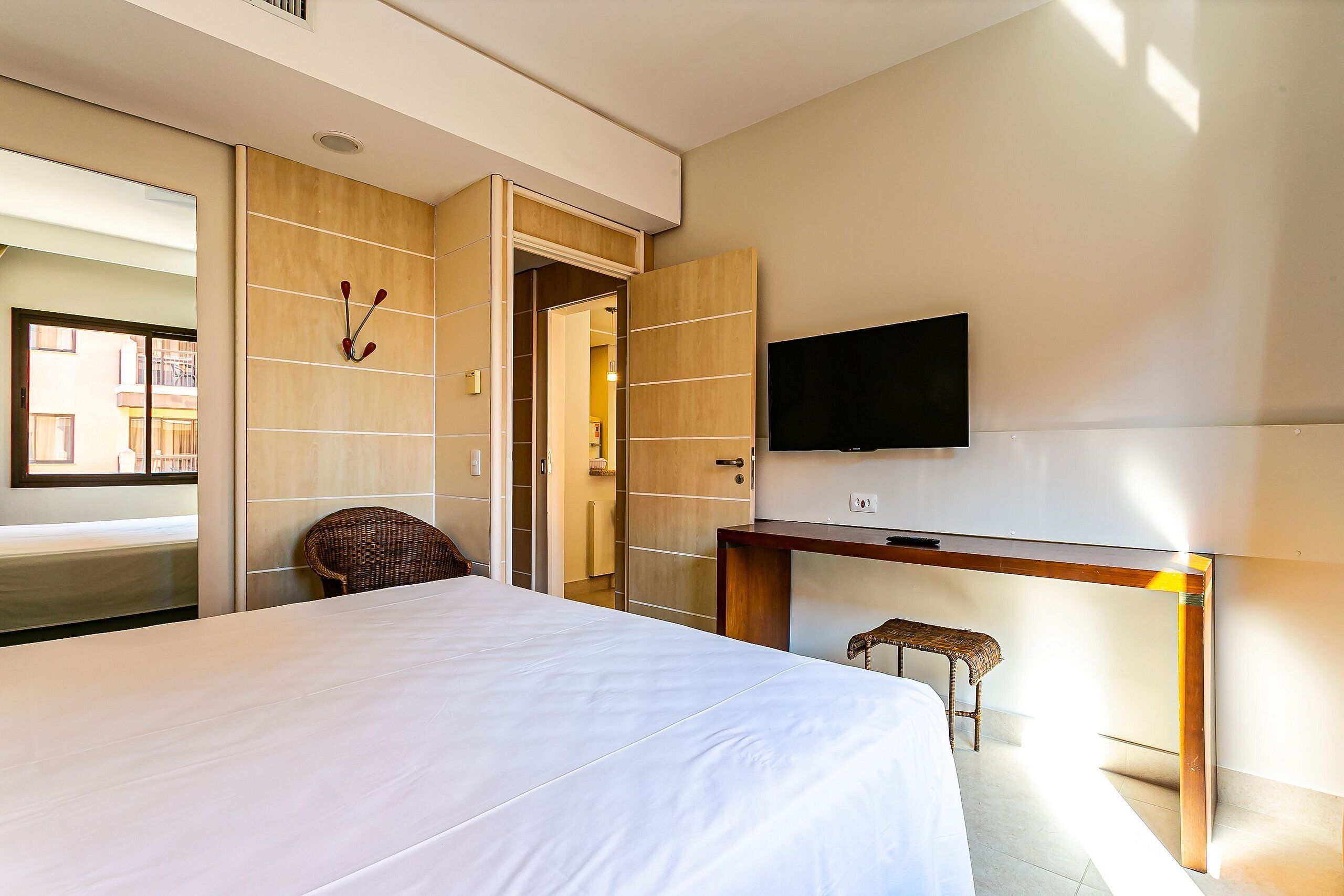 Quarto de hotel com uma cama, uma escrivaninha, uma televisão, uma cadeira e uma porta que dá para outro cômodo.