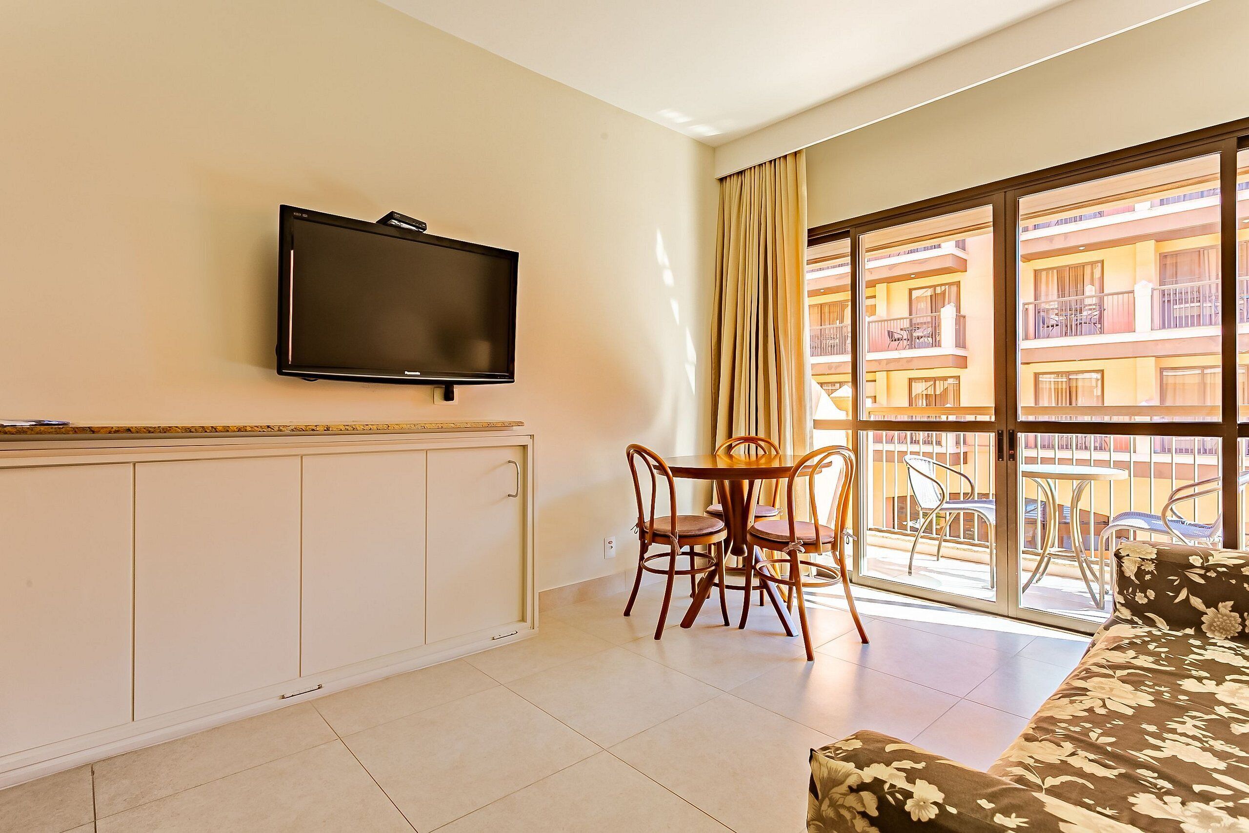 A hotel room with a TV, a dining table with chairs, a sofa, and a balcony with a view of the building.