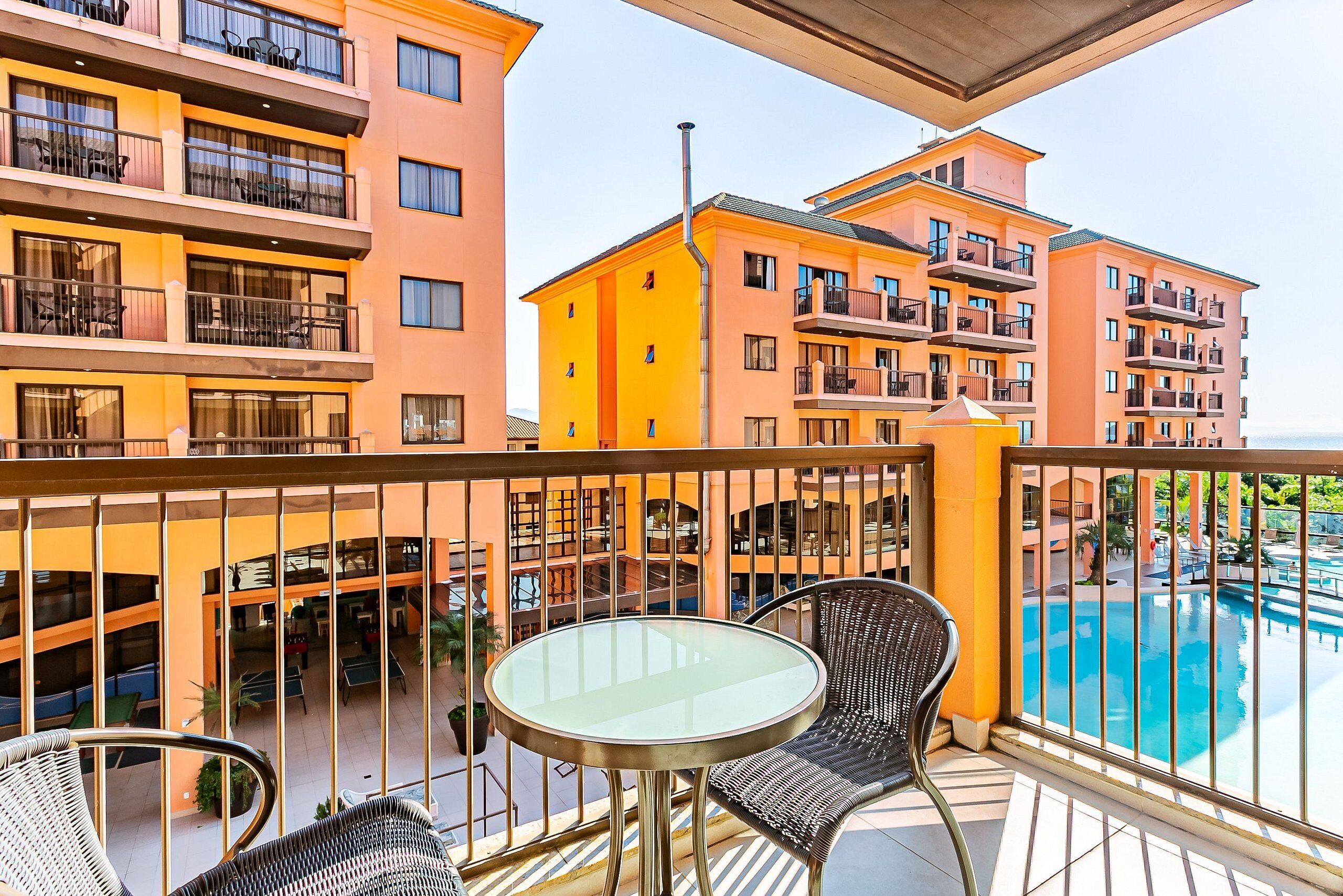Een balkon met een glazen tafel en rieten stoelen, met uitzicht op een zwembad en aangrenzende gebouwen.