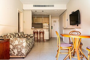A living room with a sofa, a wooden table with chairs, a kitchen area with cabinets and a TV. - Suite w/ Sea/pool View Luxury Resort Jurere Jbv142 (Florianópolis)