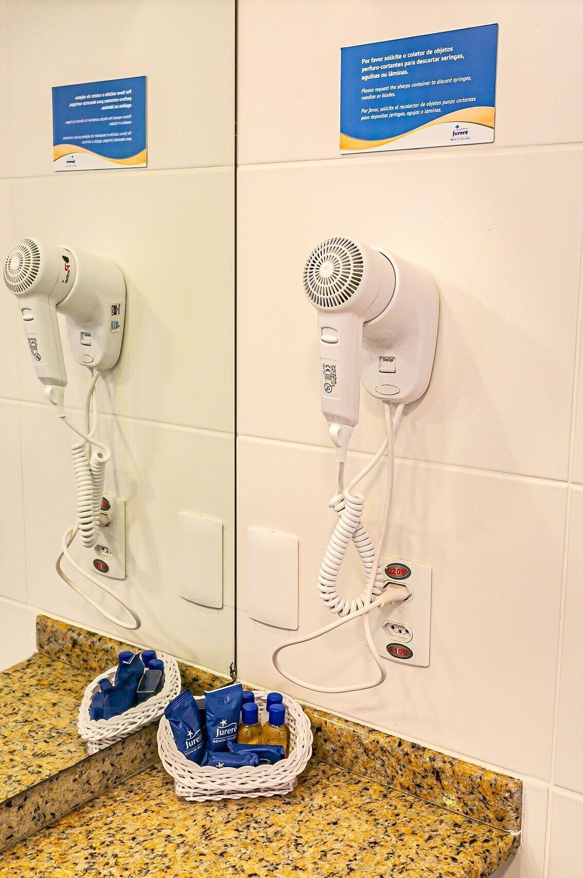 Bagno con due asciugacapelli fissati alla parete, un cesto con articoli da toilette e un cartello che invita a richiedere contenitori per oggetti taglienti da smaltire.