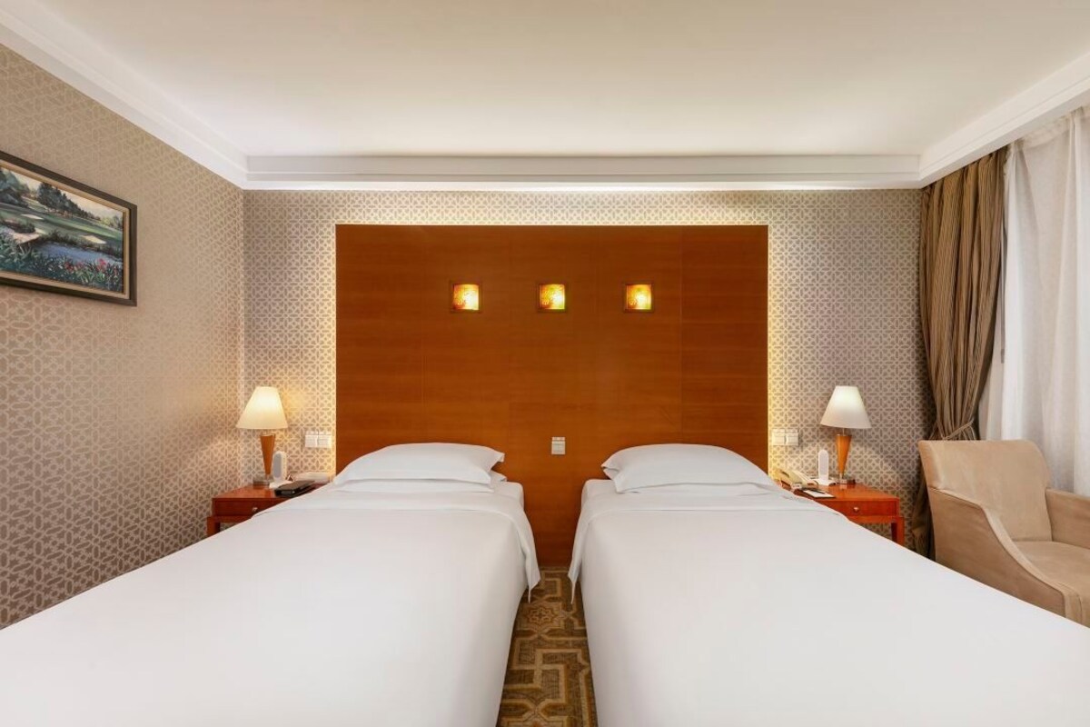 A hotel room with two beds, a wooden headboard, patterned wallpaper, a painting on the wall, and a chair.