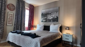 Quarto de hotel com cama, mesa de cabeceira, relógio, quadro emoldurado e cortinas.