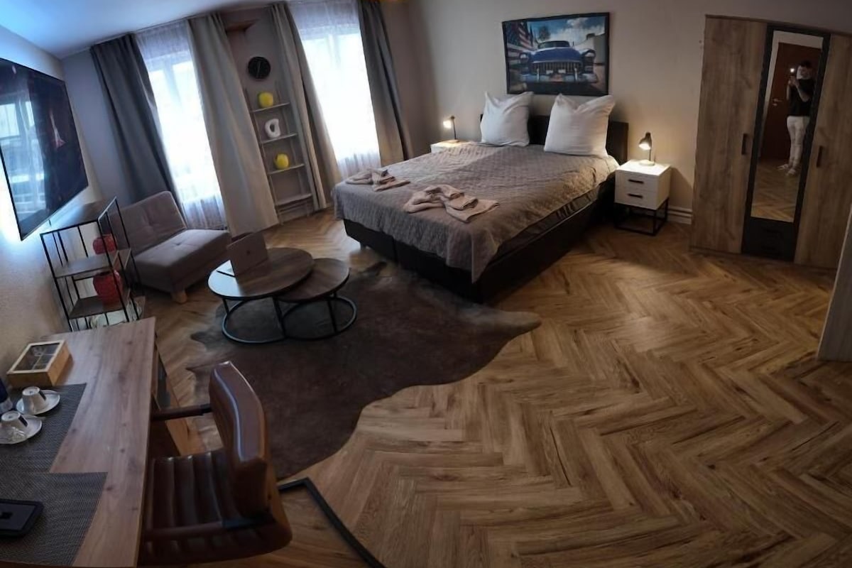Quarto de hotel com cama, escrivaninha, cadeira, mesa redonda e uma janela grande.