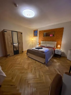 Quarto com piso de madeira em padrão herringbone, uma cama com cabeceira branca, um guarda-roupa e uma mesa de cabeceira com abajur.
