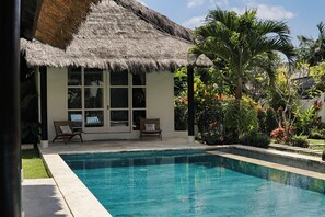 A pool area with a thatched-roof structure, two lounge chairs, and lush tropical plants. - Coastal Garden Villa (Ungasan)