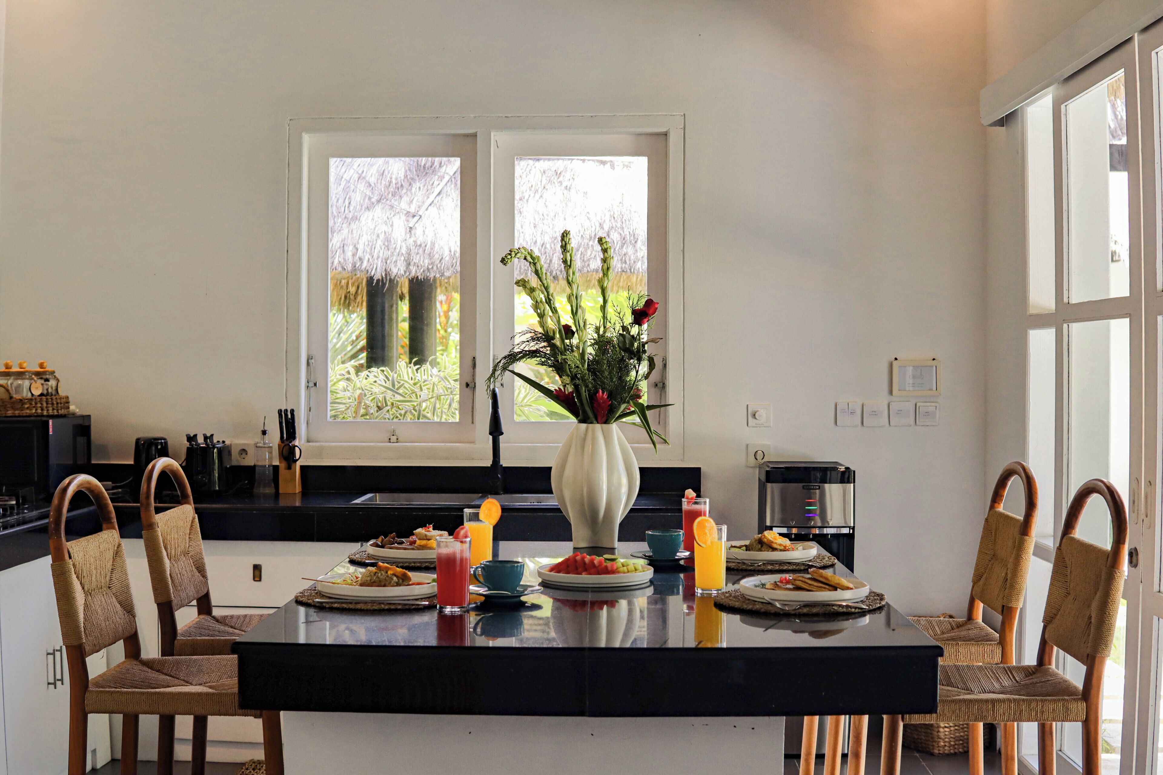 Uma cozinha moderna com uma ilha central, cadeiras de jantar, um vaso com flores e vista para o verde pela janela.
