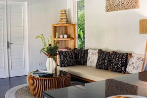 A living room with a sofa, patterned cushions, a round wooden side table, a white vase with flowers, a bookshelf, and a door. - Coastal Garden Villa (Ungasan)