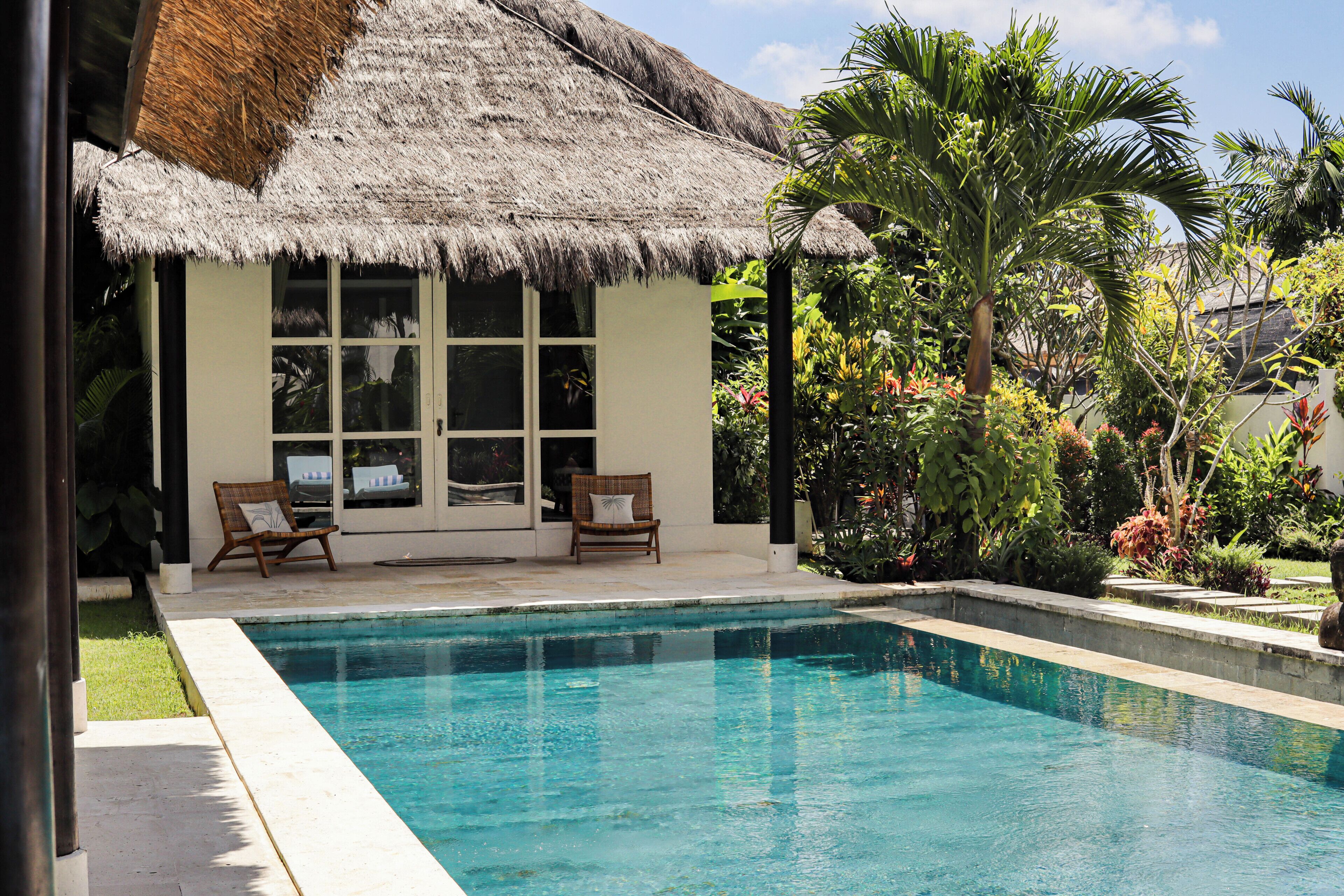 Un espace aménagé au bord de la piscine, doté d’une structure à toit de chaume, de deux chaises longues et d’une variété de plantes tropicales.