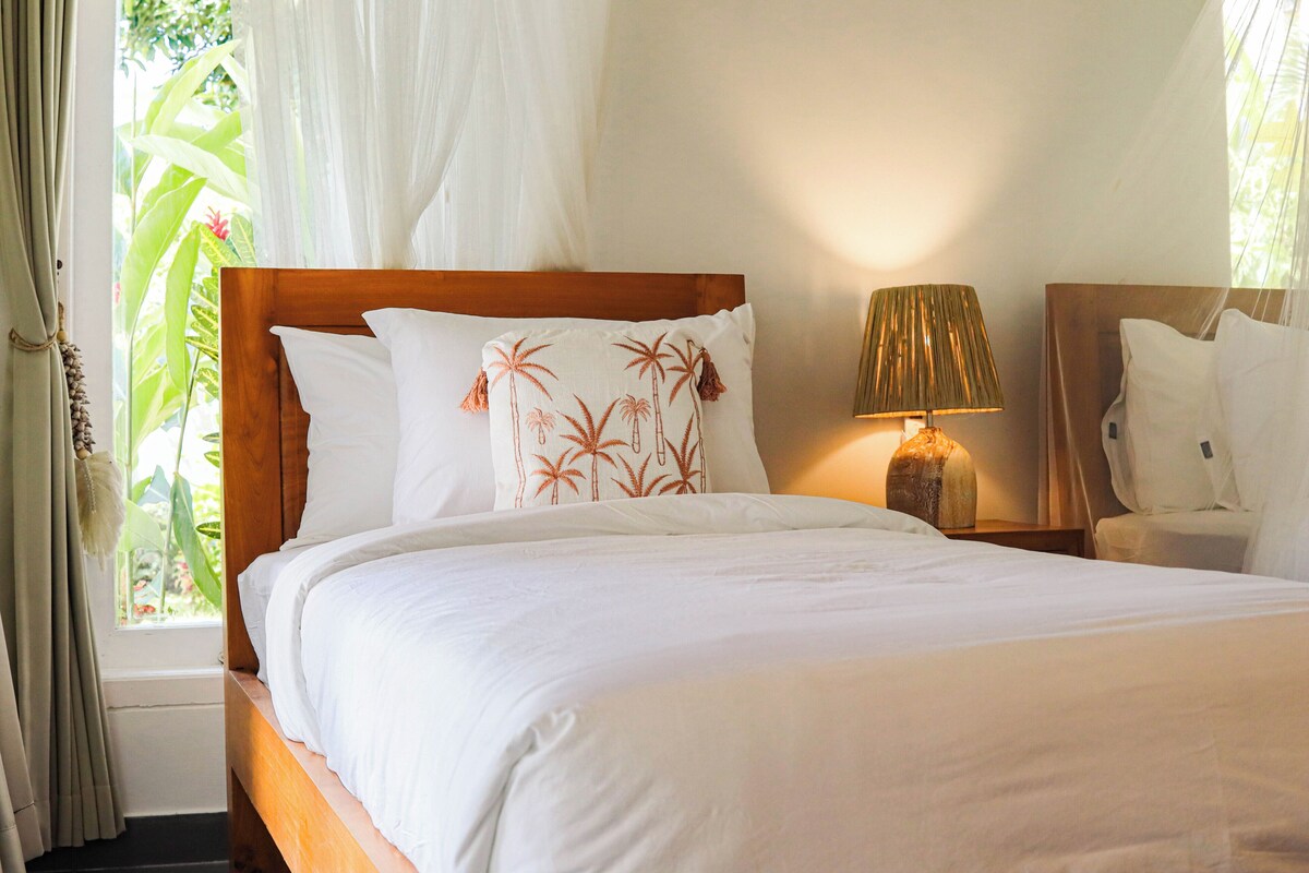 A hotel room with a wooden bed, white bedding, a nightstand with a lamp, and a view of greenery through the window.