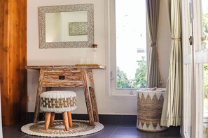 Een kamer met een houten bureau, een geweven kruk, een kleine ronde tafel, een spiegel en een raam met gordijnen.