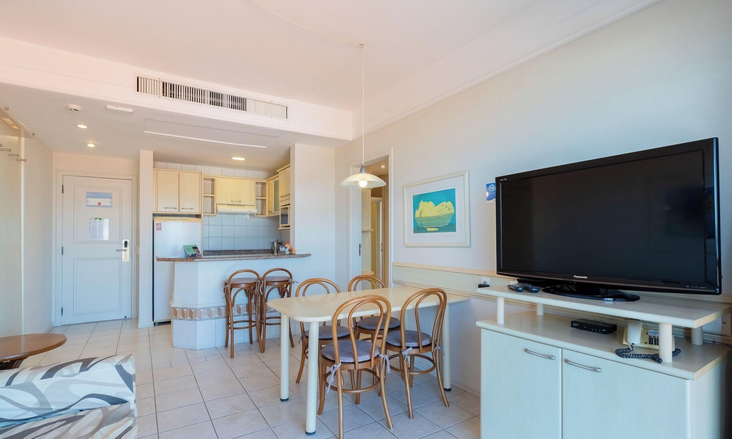 A modern living room with a dining area, a kitchen in the background, and a large flat-screen TV on a stand.