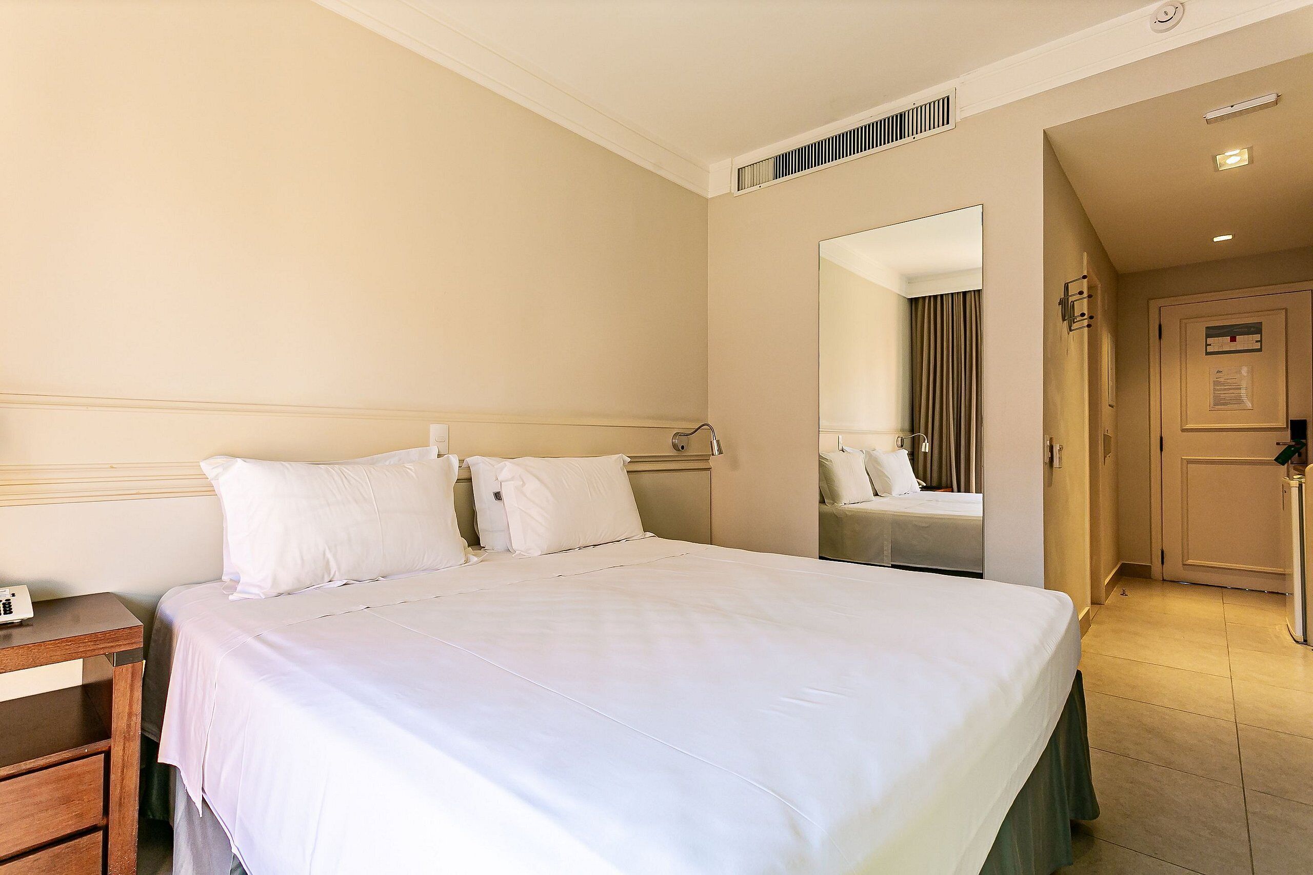 A hotel room with a large bed, a bedside table, a mirror, and a door leading to a bathroom.