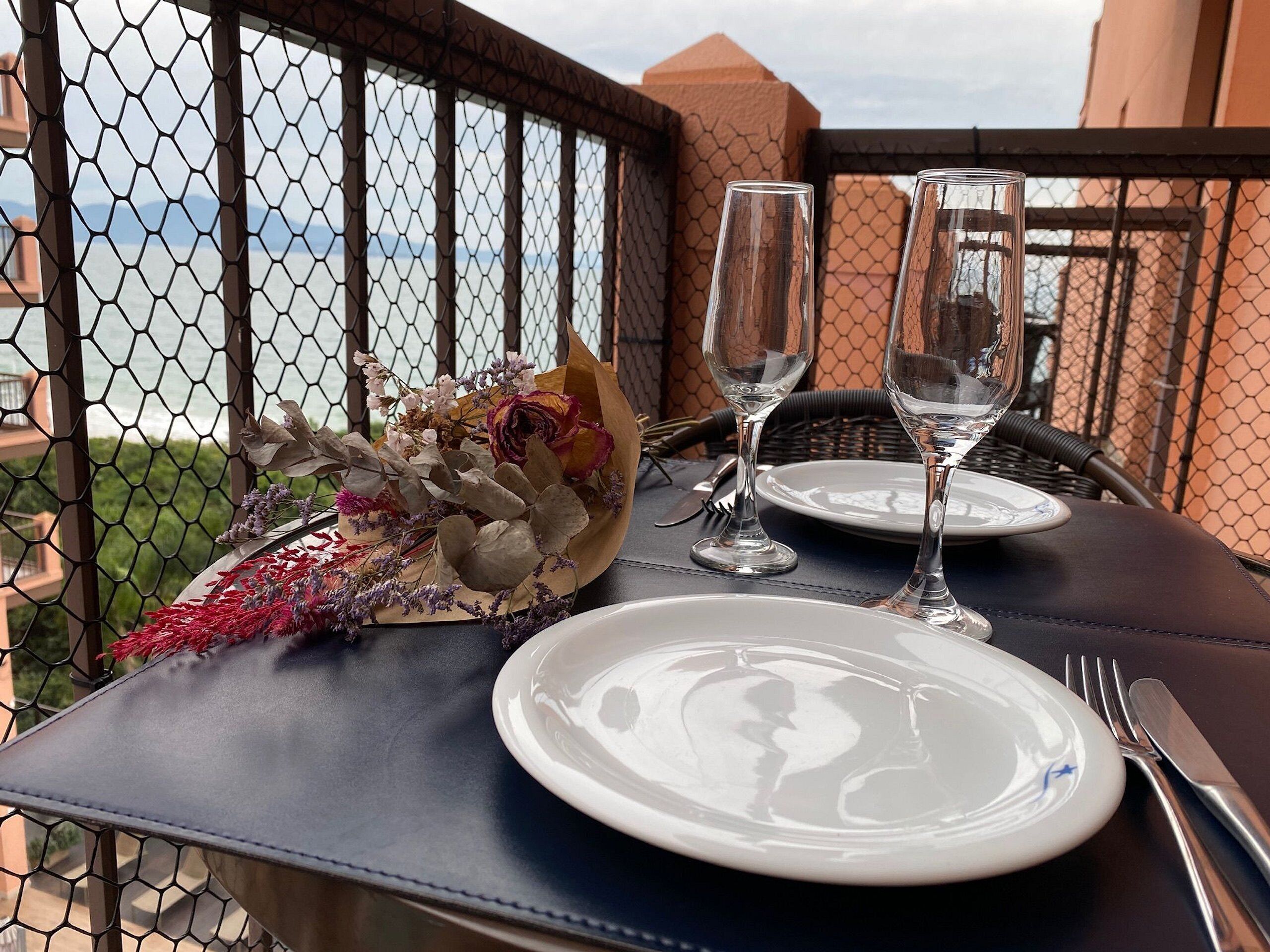Mesa para dos, con un ramo de flores, dos copas de vino y un plato en un balcón con vista al mar.