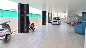 A covered parking area with a scooter and a car parked, a blue seating area, and a building in the background. - HOTEL SIDDVEER GRAND (Guntur)