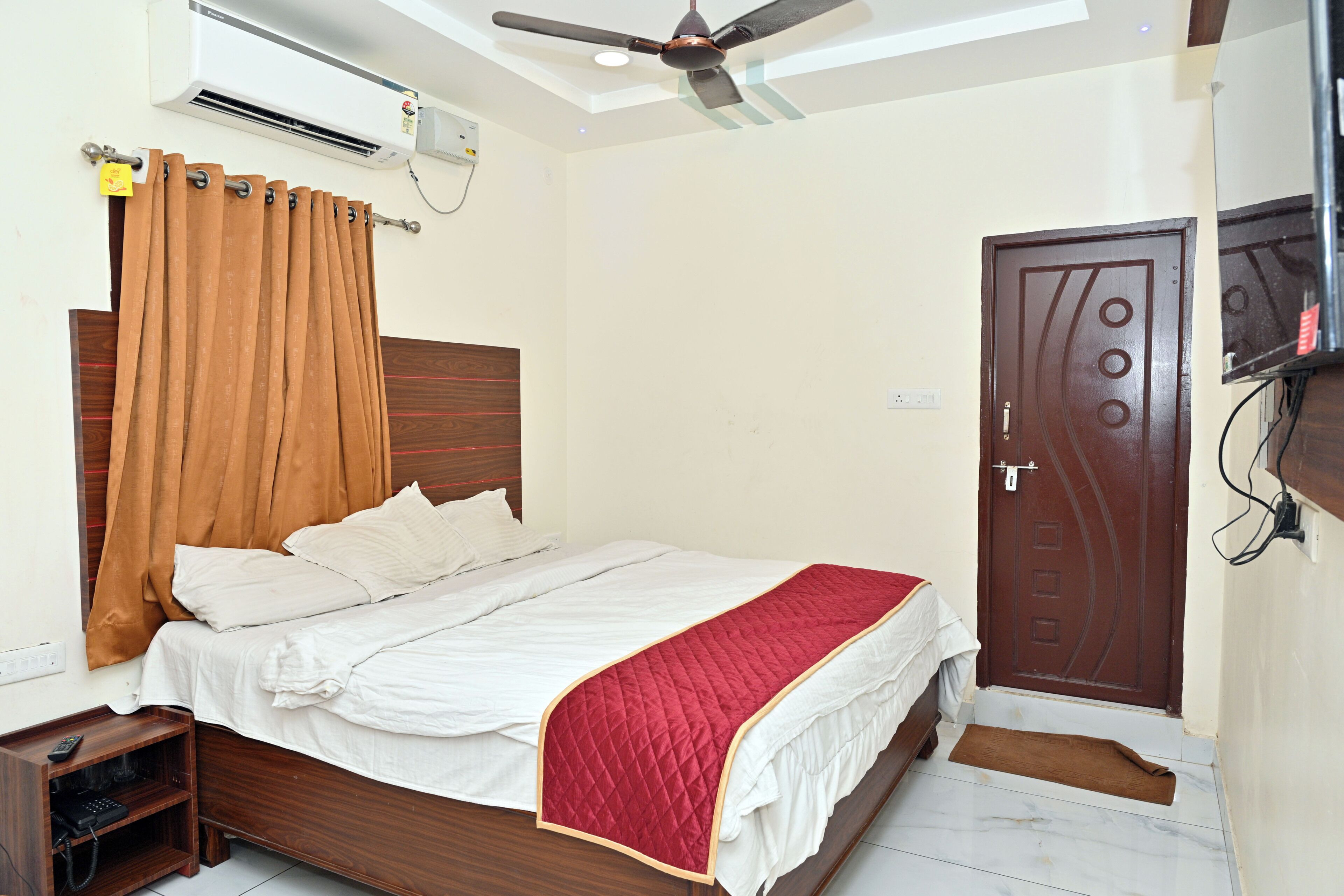 A bedroom with a bed, a ceiling fan, an air conditioning unit, a wooden nightstand, and a door.