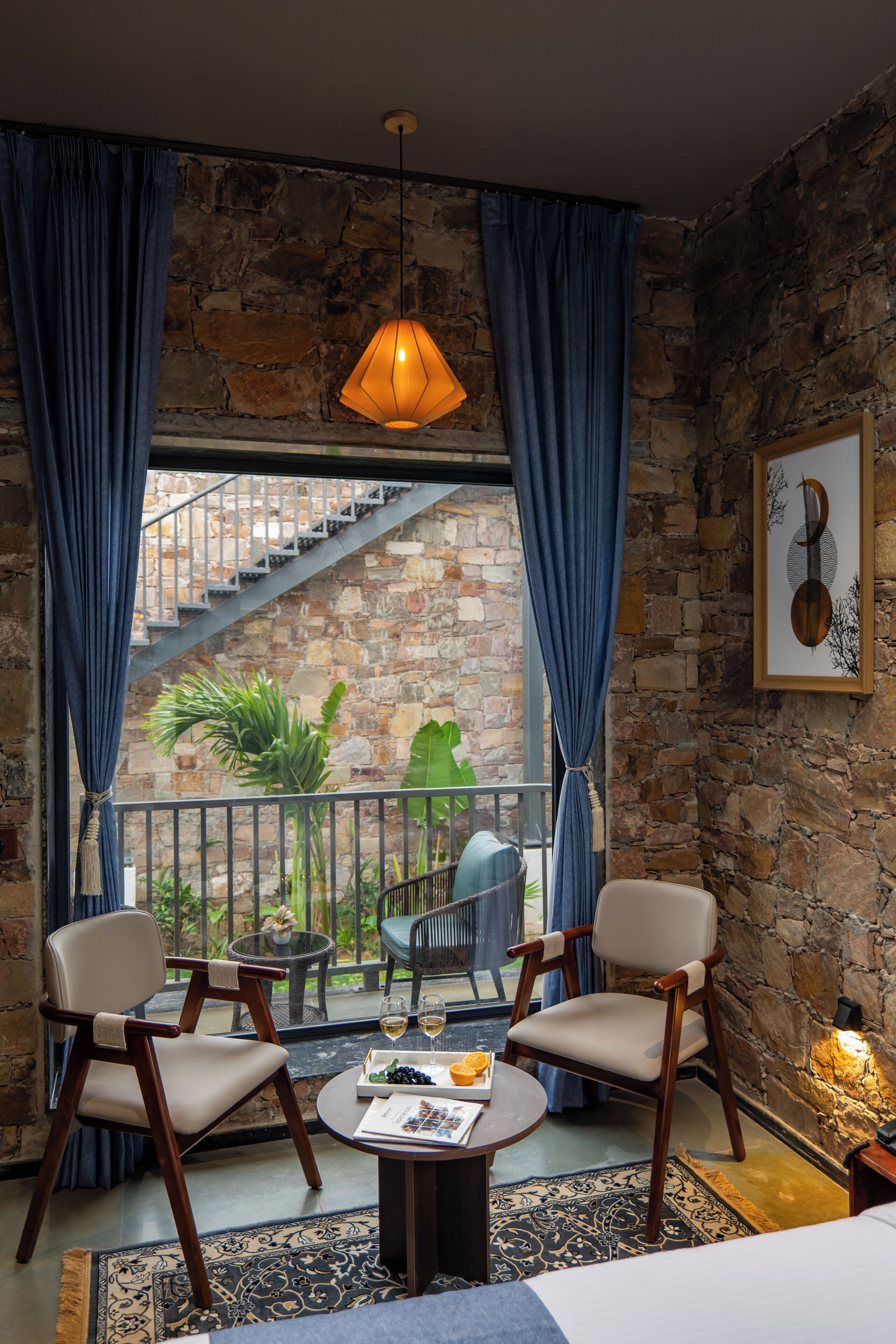 Une pièce avec un mur en pierre, un escalier, deux chaises, une table sur laquelle se trouve un plateau de fruits et des verres, et un luminaire suspendu.