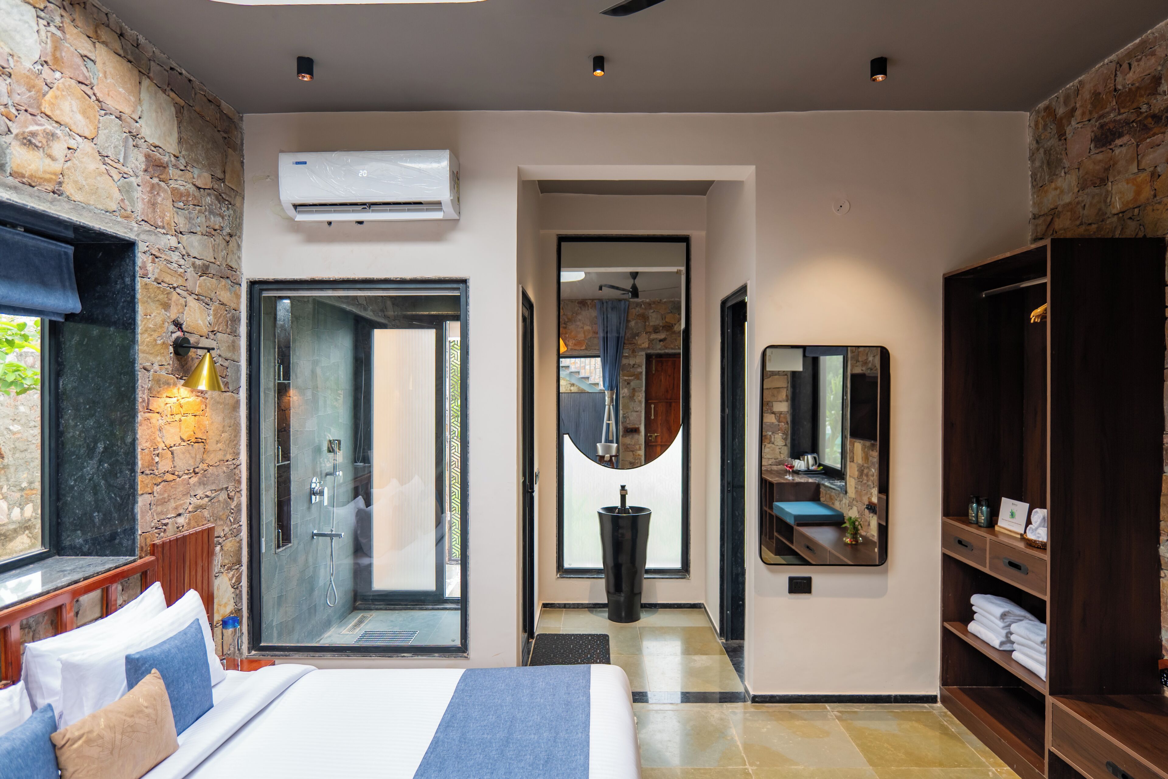 A hotel room with a stone wall, a large bed, a bathroom with a glass door, and a mirror reflecting the room.