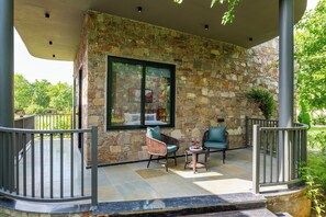 A stone exterior wall with a large window, outdoor seating with cushions, and a metal railing.