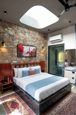 A modern hotel room with a stone wall, a large bed with a blue and white bedspread, a wooden headboard, a nightstand, and a glass shower enclosure.