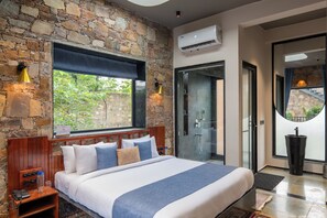 A bedroom with a stone wall, a large window, a bed with multiple pillows, a bedside table, a telephone, and an air conditioning unit. - Hathnoda Bagh Cottage ELIVAAS Signature (Jaipur)