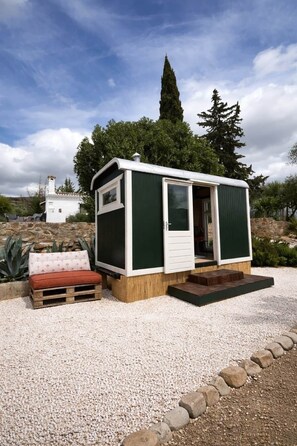 A small, single-story mobile home with a white door and a window, set on a gravel surface with a stone border, surrounded by greenery and a clear sky.