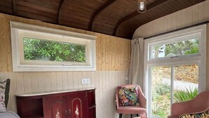 Una habitación con techo de madera, una cama con ropa de cama floral, un mueble de madera oscura, una silla rosa con cojín floral, y una ventana con vistas a zonas verdes.