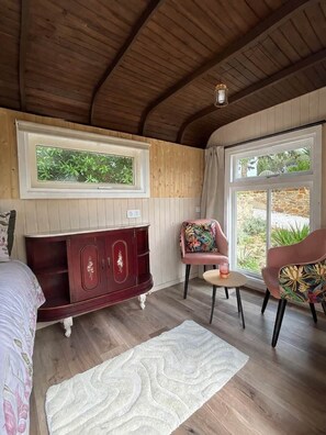 A room with a wooden ceiling, a bed with floral bedding, a dark wooden cabinet, a pink chair with a floral cushion, and a window with a view of greenery.