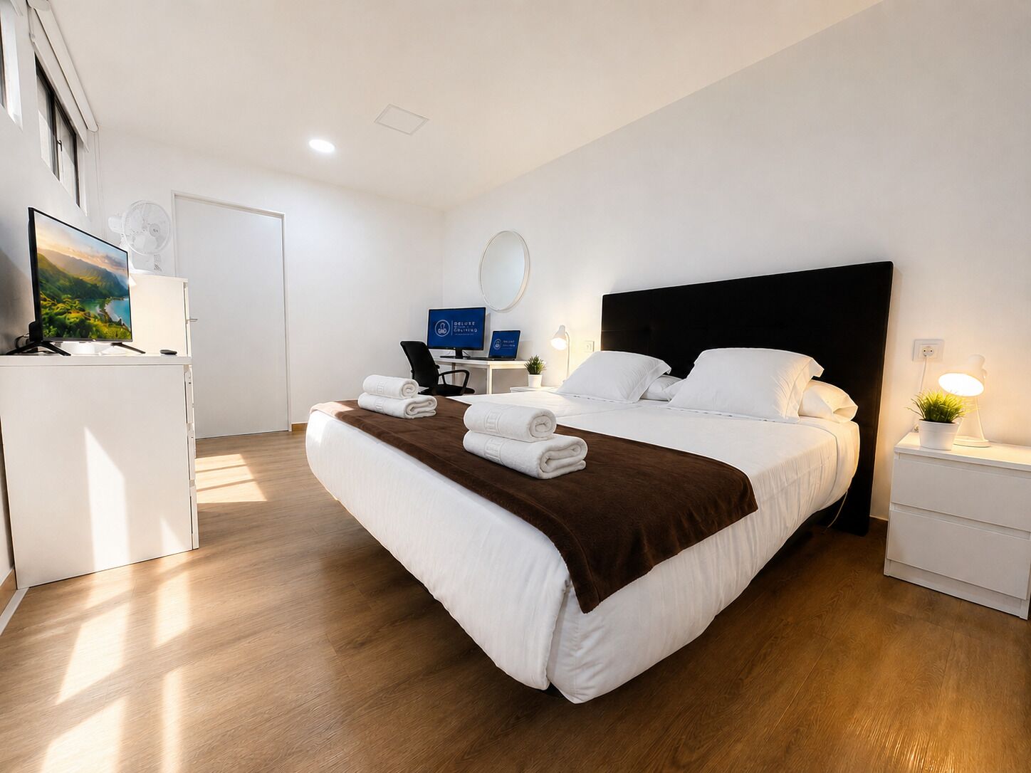 A hotel room with a bed, a desk with a computer, a TV, and a bedside lamp.