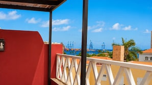A balcony with a white railing, overlooking a port with cranes and ships, and a red building in the foreground.