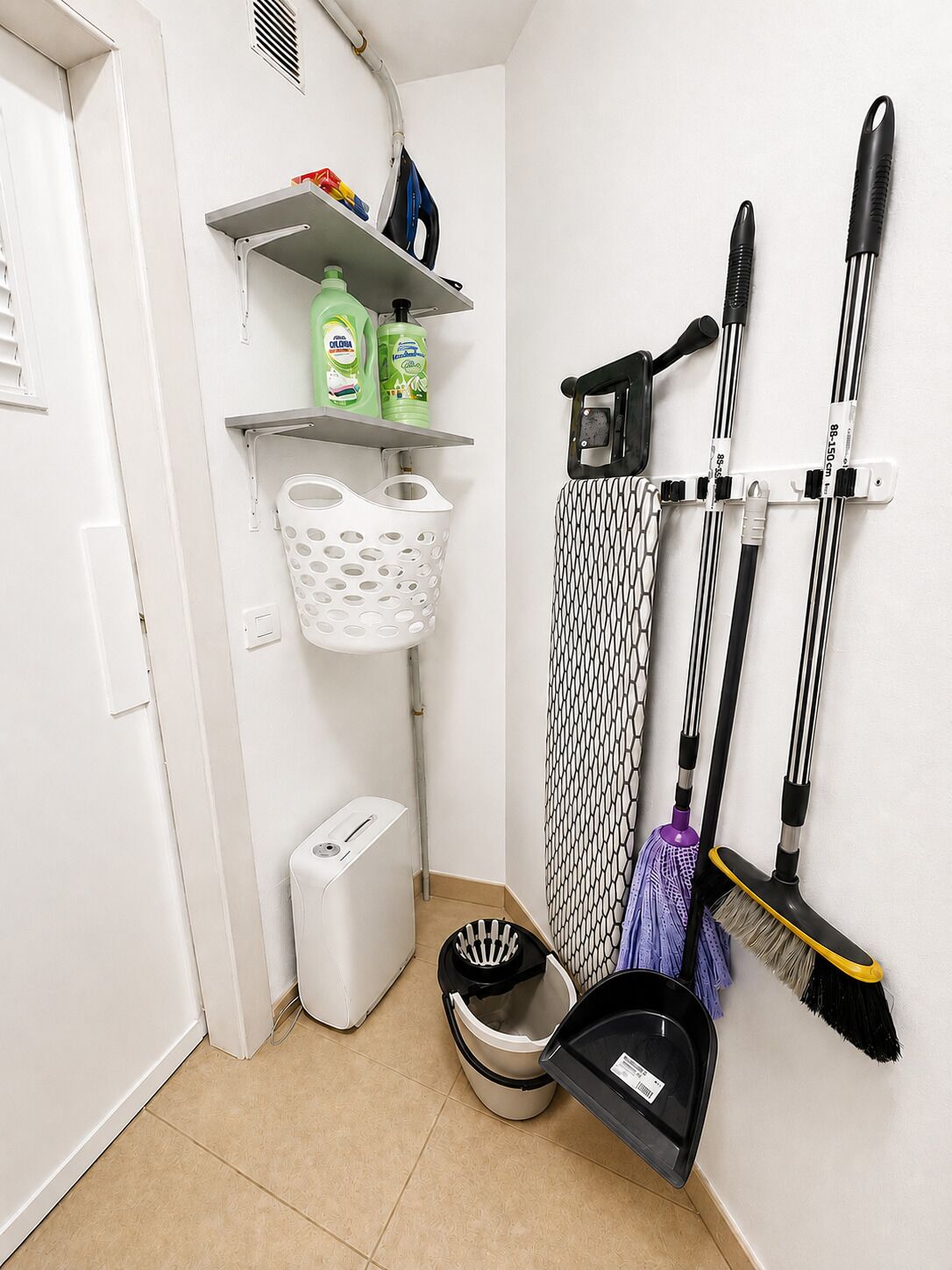 Esquina de una habitación con un trapeador, escoba, cesta de ropa y productos de limpieza en un estante adosado a la pared.