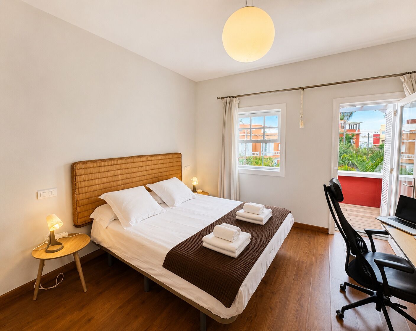 A hotel room with a bed, bedside lamp, desk with chair, and a view of buildings through the window.